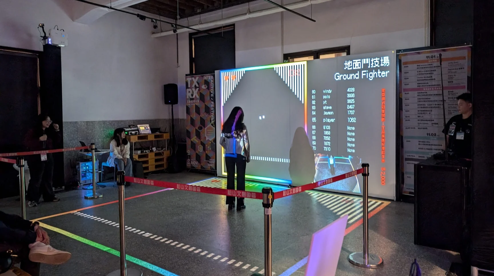地面鬥技場的現場照片：昏暗展場裡一面發光的投影牆顯示遊戲畫面與排行榜，一名玩家背對鏡頭站在地面投影的互動區裡，四周以紅色伸縮帶圍欄圍出範圍，左側是控制桌與工作人員。